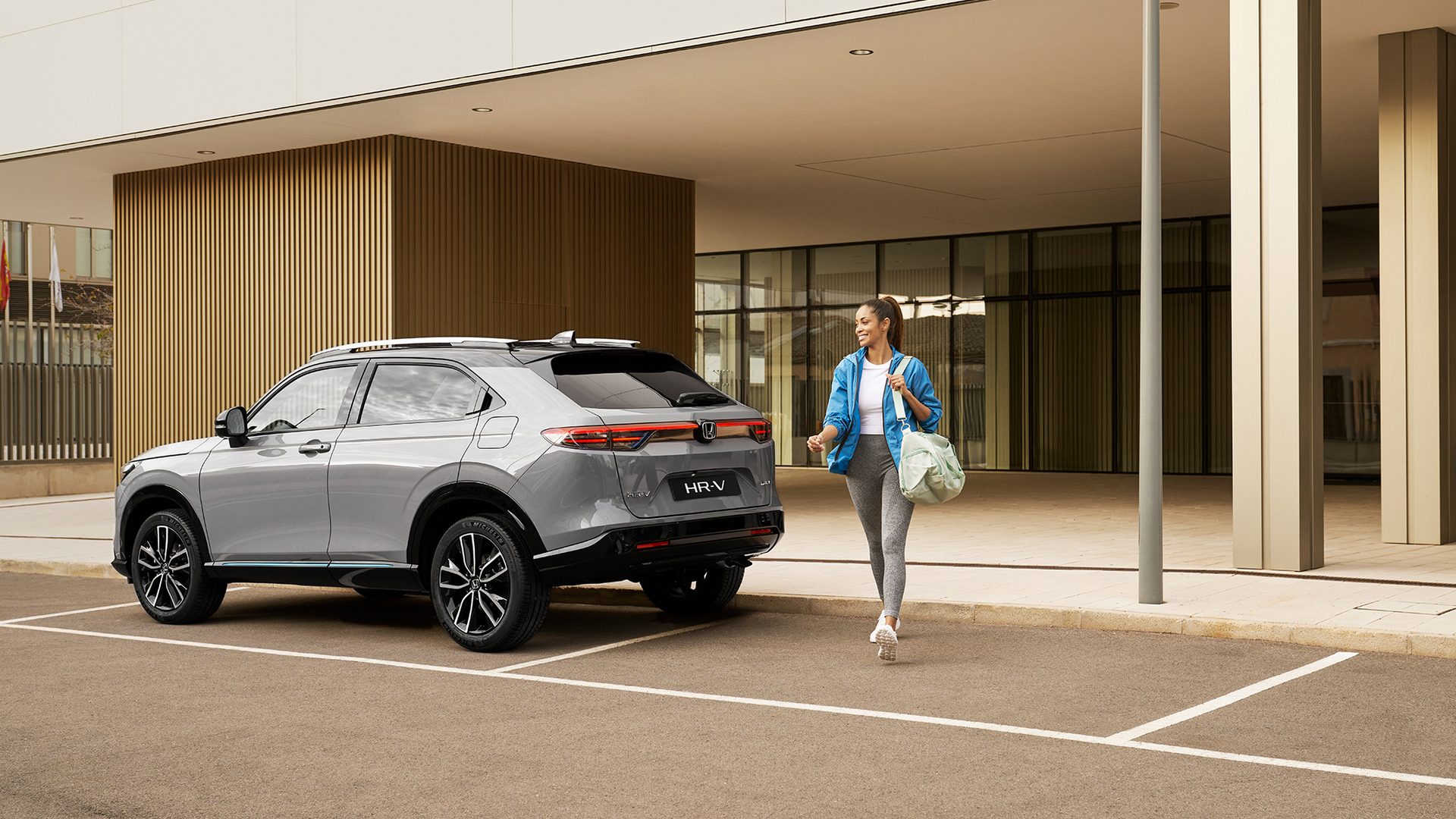 Strieborná Honda HR-V zaparkovaná pred modernou budovou, zatiaľ čo okolo nej prechádza žena s taškou, čo naznačuje každodenné mestské využitie a komfort.