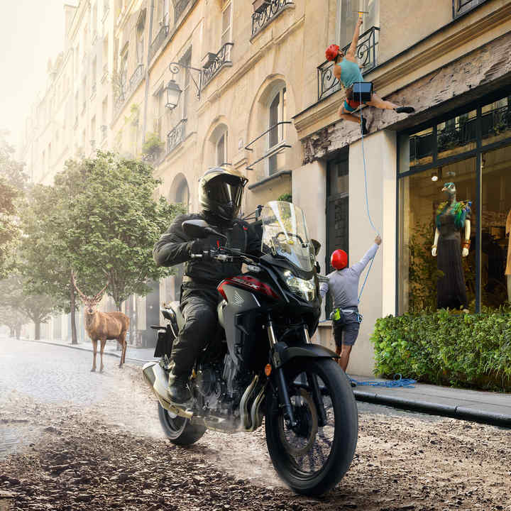Trojštvrťový pohľad spredu z ľavej strany na model CB500X s jazdcom pri jazde po znečistenej mestskej ulici 