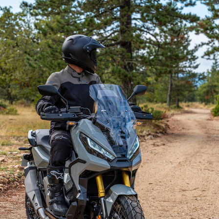 Honda X-ADV, trojštvrťový predný záber z pravej strany, jazdec stojaci na sivom motocykli, lesná cesta