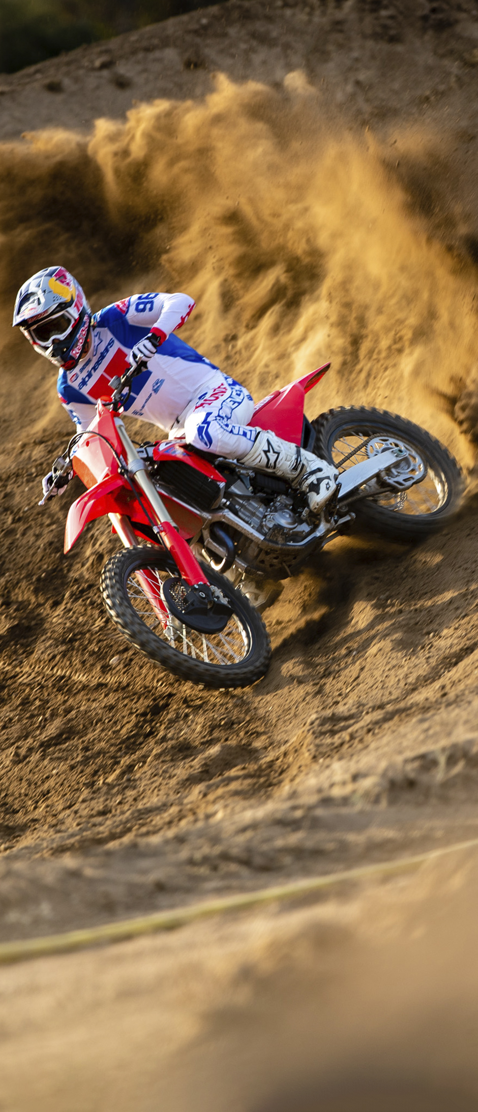 Motokrosový jazdec v bielom, modrom a červenom výstroji nakláňa červenú motokrosovú motorku do strmého pieskového valu, pričom vyhadzuje do vzduchu veľkú vlnu zeminy pri jazde zákrutou.