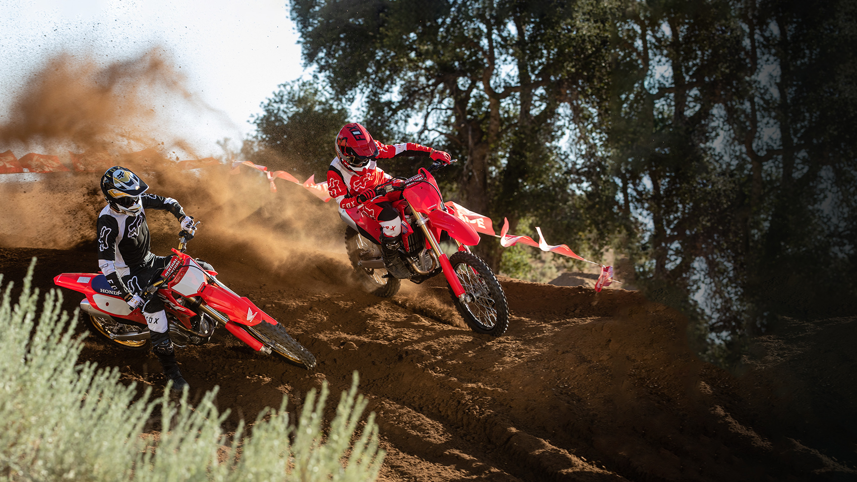 Two motocross riders in full protective gear race red dirt bikes around a dusty off-road track, kicking up clouds of dirt as they lean sharply into a turn; trees and course tape line the background, and a small banner reads “Off Road — Let the fun begin.lder 
