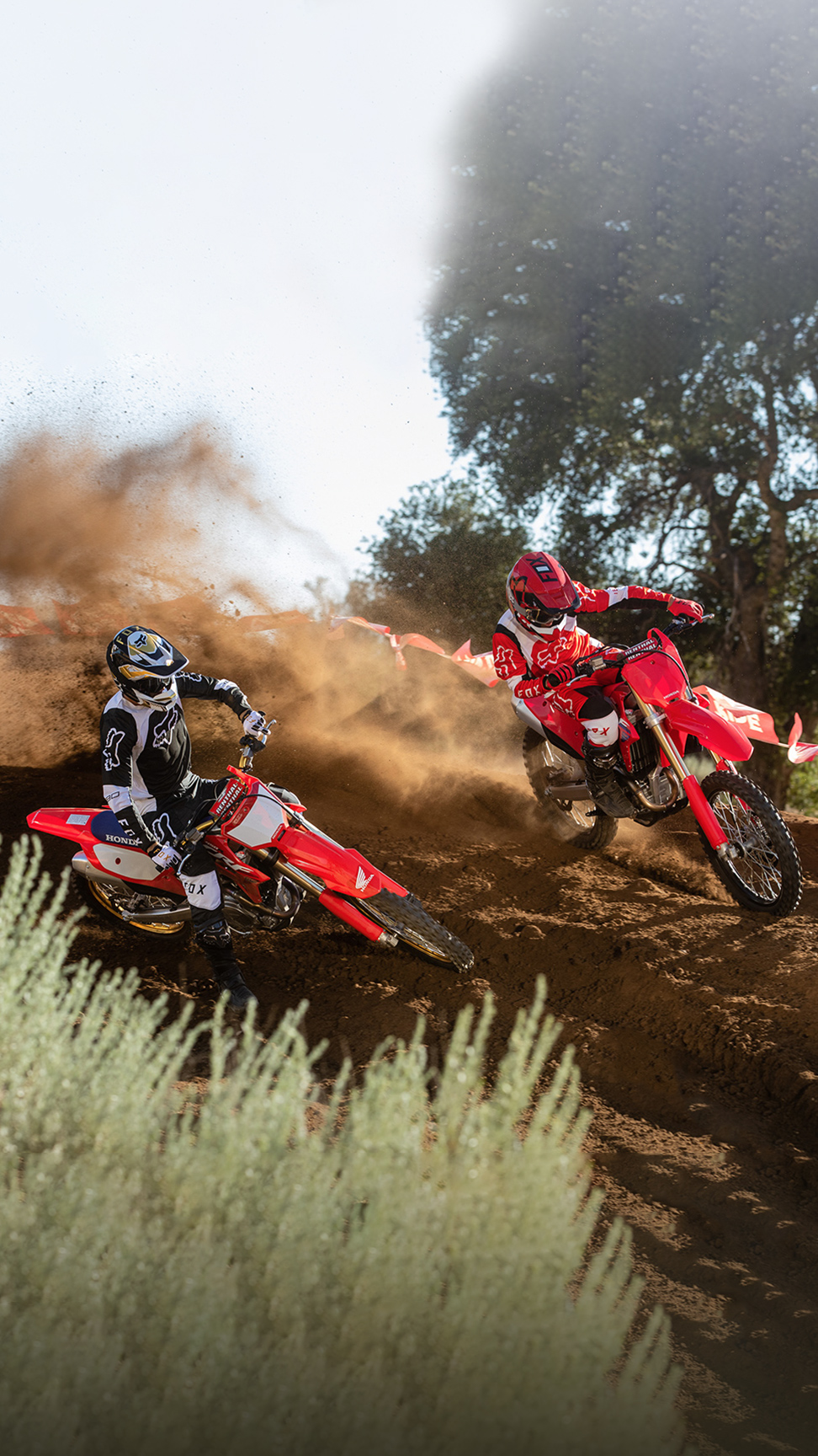 Two motocross riders in full protective gear race red dirt bikes around a dusty off-road track, kicking up clouds of dirt as they lean sharply into a turn; trees and course tape line the background, and a small banner reads “Off Road — Let the fun begin.lder 