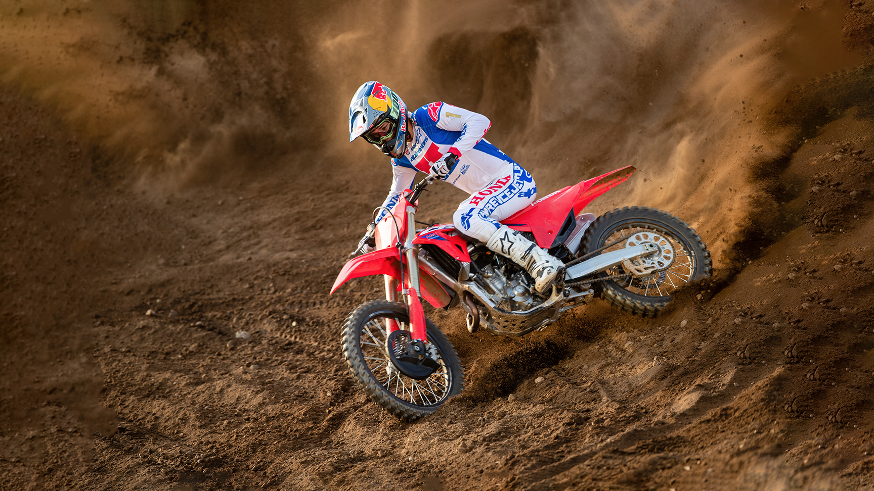 Motokrosový jazdec v bielom, modrom a červenom pretekárskom výstroji sa ostro nakláňa do zákruty na červenej motokrosovej motorke, prereže piesočnú trať a za sebou zanecháva pôsobivý oblak piesku.