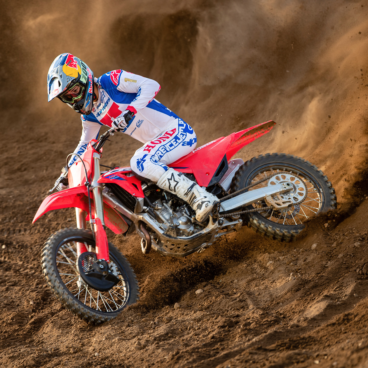 Motokrosový jazdec v bielom, modrom a červenom pretekárskom výstroji sa ostro nakláňa do zákruty na červenej motokrosovej motorke, prereže piesočnú trať a za sebou zanecháva pôsobivý oblak piesku.