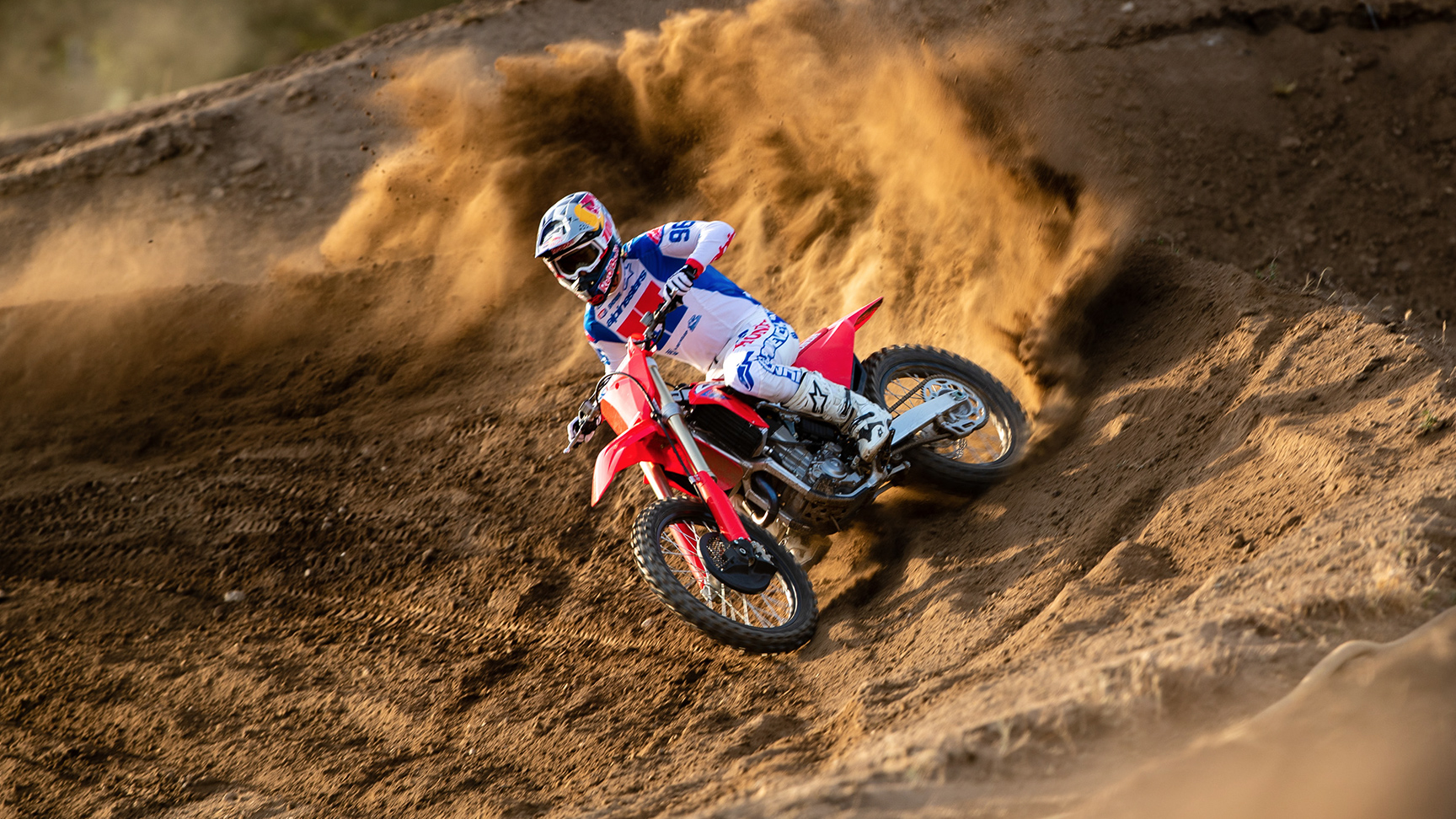 Motokrosový jazdec v bielom, modrom a červenom výstroji nakláňa červenú motokrosovú motorku do strmého pieskového valu, pričom vyhadzuje do vzduchu veľkú vlnu zeminy pri jazde zákrutou.